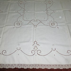 K&ouml;nnte beinhalten: Wei&szlig;e bestickte Tischdecke mit zarter Spitzenbord&uuml;re. Die Tischdecke zeigt ein sich wiederholendes Muster aus floralen Motiven und filigranen Schn&ouml;rkeln.