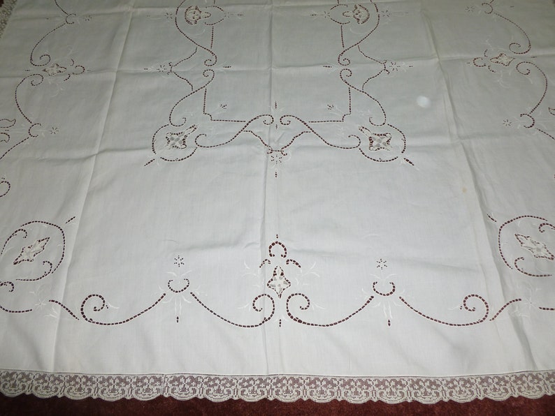 K&ouml;nnte beinhalten: Wei&szlig;e bestickte Tischdecke mit zarter Spitzenbord&uuml;re. Die Tischdecke zeigt ein sich wiederholendes Muster aus floralen Motiven und filigranen Schn&ouml;rkeln.