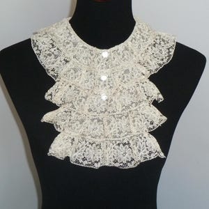 May include: Cream-colored lace collar with three layers of ruffles and small, round buttons. The collar is displayed on a black mannequin, showcasing its delicate floral pattern and vintage style.