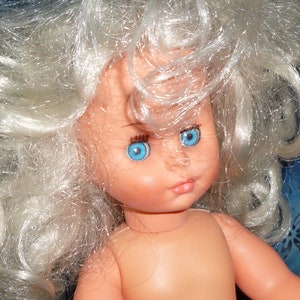 Puede incluir: Una mu&ntilde;eca con el pelo largo, blanco y rizado y ojos azules. La mu&ntilde;eca no lleva ropa y tiene una tez rosa claro.