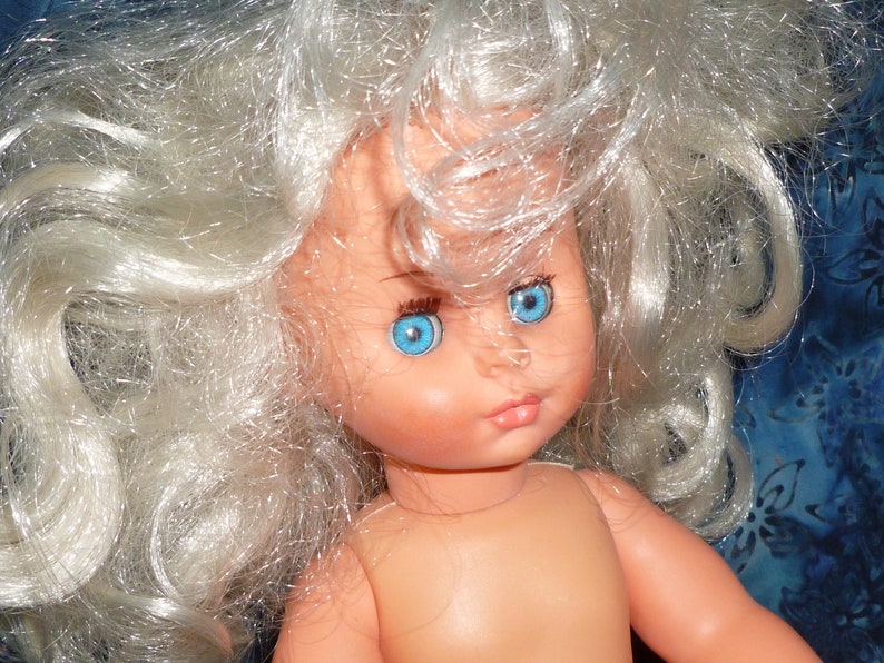 Puede incluir: Una mu&ntilde;eca con el pelo largo, blanco y rizado y ojos azules. La mu&ntilde;eca no lleva ropa y tiene una tez rosa claro.