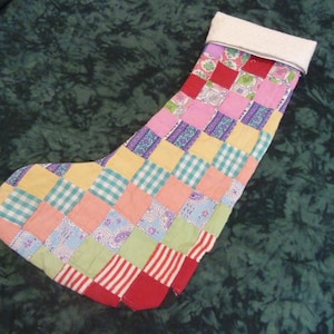 May include: A patchwork Christmas stocking in various colours. The design includes squares of pink, yellow, blue, and green fabric. The cuff is white, and the toe is red with white stripes. A festive, handmade item.