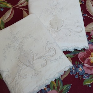 May include: Two white embroidered pillowcases with a floral design. The pillowcases feature a detailed embroidered pattern of a vase and floral elements. The edges are scalloped. The background is a floral patterned fabric in shades of red, pink, and blue.