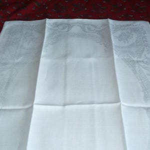 Op de afbeelding: Wit linnen tafelkleed met een delicaat geborduurd patroon van bladeren en bloemen. Het tafelkleed is in drie&euml;n gevouwen, waardoor het patroon aan alle drie kanten zichtbaar is.