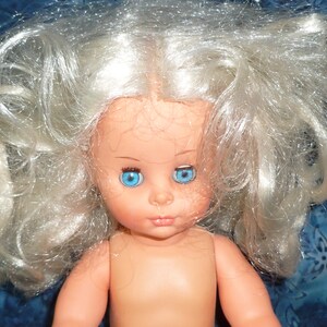 Puede incluir: Una mu&ntilde;eca con el pelo largo, blanco y rizado y ojos azules. La mu&ntilde;eca no lleva ropa y tiene una tez rosa claro.