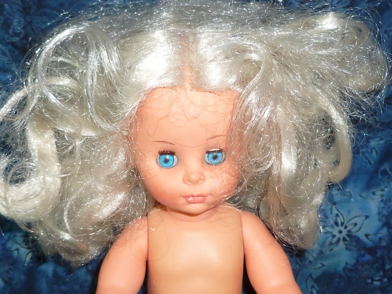 Puede incluir: Una mu&ntilde;eca con el pelo largo, blanco y rizado y ojos azules. La mu&ntilde;eca no lleva ropa y tiene una tez rosa claro.