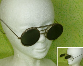 May include: A pair of vintage, round, dark-tinted sunglasses with a silver metal frame. The sunglasses have a unique design with a hinged, flip-up feature.