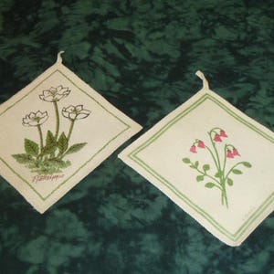 May include: Two off-white, diamond-shaped pot holders with floral designs. One features white flowers with green leaves and the word "Frällsippe". The other has pink bell-shaped flowers and green leaves. Both have green borders.