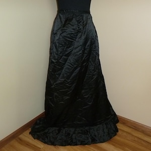 May include: A long, black satin skirt with a ruffled hem. The skirt has a high waist and a flowing silhouette. The fabric appears to be slightly wrinkled, adding texture. The skirt is suitable for formal occasions or costume events.