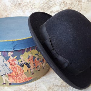 Może przedstawiać: Czarny filcowy kapelusz fedora z czarną wstążką grosgrain. Kapelusz leży na okrągłym pudełku na kapelusze z niebieskim i białym wzorem.