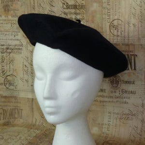 May include: A black beret hat, a classic French style, is displayed on a white mannequin head. The hat is made of a soft, dark material and has a round shape. The background features vintage-style paper.
