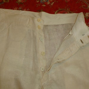 May include: A pair of white linen pants with a button fly closure. The pants have a waistband with five buttons.