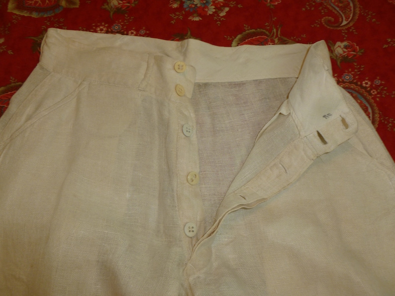 May include: A pair of white linen pants with a button fly closure. The pants have a waistband with five buttons.