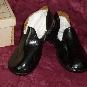 Peut inclure: Une paire de surchaussures en caoutchouc noir avec une doublure en papier blanc à l'intérieur. Les surchaussures sont posées sur une surface en tissu bordeaux. La boîte des surchaussures est visible en arrière-plan. La boîte est blanche avec du texte brun qui dit "RALL-BAND Co., Mishawaka, Indiana".