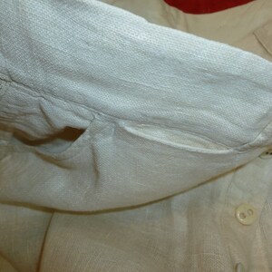 May include: A white linen shirt with a pocket and a button closure. The shirt is folded over and the pocket is visible.