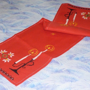 May include: Red table runner with white embroidered snowflakes and candles. The runner is folded in half and has a white stitched border.