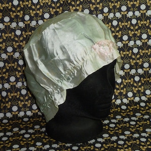 May include: A light green satin sleep cap with a pink flower embellishment. The cap is on a black mannequin head.