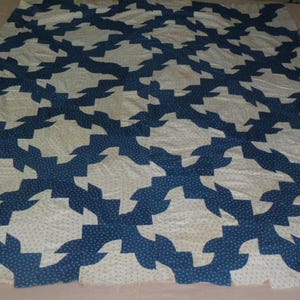 May include: A vintage quilt with a repeating pattern of white shapes against a dark blue background. The quilt is made of fabric with a dotted pattern. The overall design is symmetrical and geometric, creating a visually appealing textile.