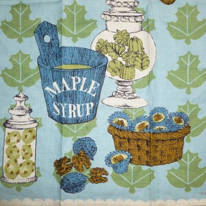 May include: A blue and white kitchen towel with a maple syrup bucket, a jar of nuts, a bowl of candies, and green maple leaves. The towel has the text "Maple Syrup" on the bucket.