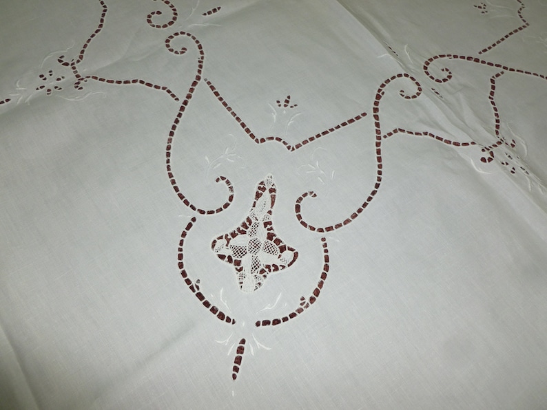 K&ouml;nnte beinhalten: Wei&szlig;e Tischdecke mit aufwendigen Stickereien aus Spitze in einem zarten Blumenmuster. Die Stickerei ist mit braunem Garn ausgef&uuml;hrt und erzeugt einen dezenten Kontrast zum wei&szlig;en Stoff.