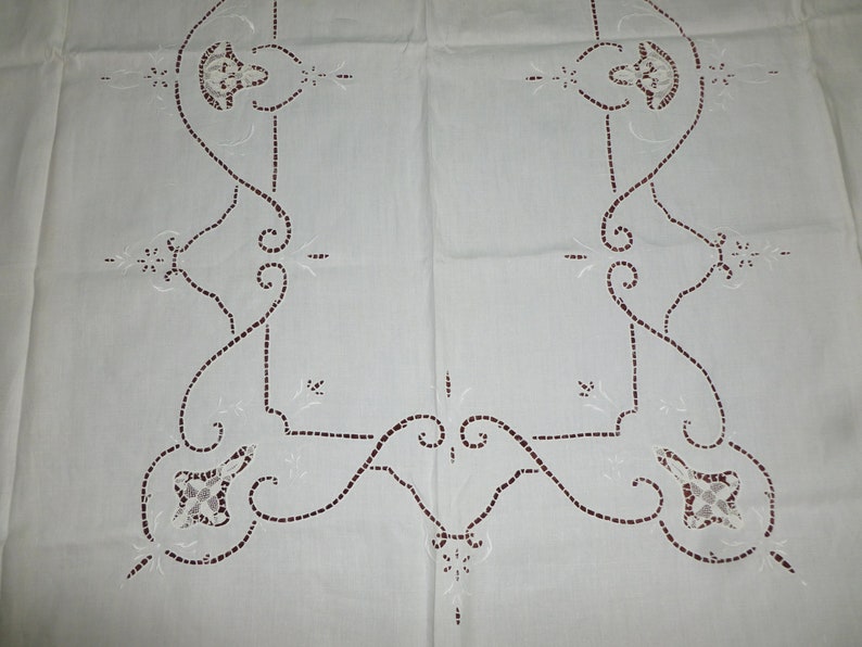 K&ouml;nnte beinhalten: Wei&szlig;e Tischdecke aus Leinen mit einer zarten Stickerei an der Kante. Die Kante zeigt ein sich wiederholendes Muster aus stilisierten Blumenmotiven in Burgunderrot.