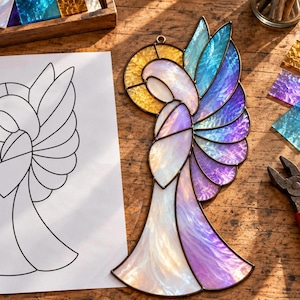 May include: A stained glass angel ornament featuring iridescent white, purple, and blue glass segments with a textured yellow halo. The piece is constructed with dark metal solder lines and includes a small metal loop at the top for hanging.
