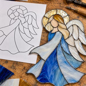 May include: A stained glass angel ornament featuring a halo of amber glass, a head of pale peach glass, and a flowing gown composed of various shades of blue and white swirled glass. The wings are crafted from white opalescent glass pieces.