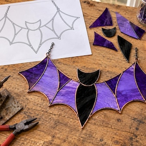 May include: A stained glass bat suncatcher featuring purple and black glass segments joined by copper foil. The bat has a central black body and wings with purple panels. A paper pattern, loose glass shards, and tools are visible nearby.