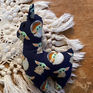 K&ouml;nnte beinhalten: Ein Stofftier in Form eines Lamas aus marineblauem Stoff, der mit Bildern einer gr&uuml;n geh&auml;uteten Figur bedruckt ist. Das Lama hat ein wei&szlig;es Gesicht und Ohren. Es steht auf einer cremefarbenen Oberfl&auml;che im Makramee-Stil.