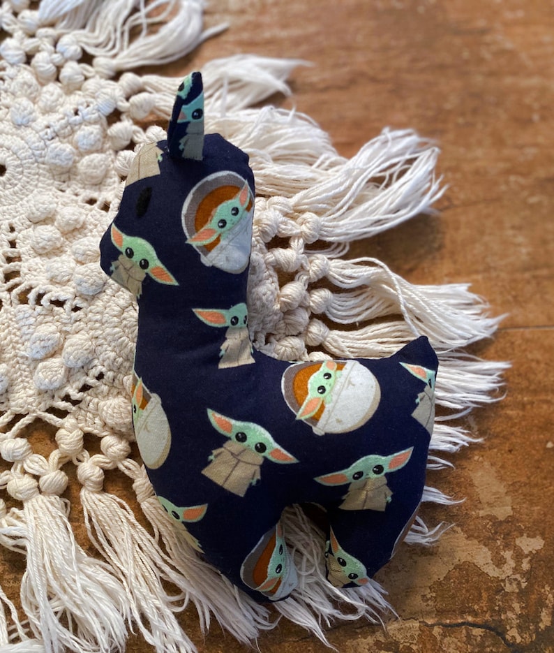 K&ouml;nnte beinhalten: Ein Stofftier in Form eines Lamas aus marineblauem Stoff, der mit Bildern einer gr&uuml;n geh&auml;uteten Figur bedruckt ist. Das Lama hat ein wei&szlig;es Gesicht und Ohren. Es steht auf einer cremefarbenen Oberfl&auml;che im Makramee-Stil.