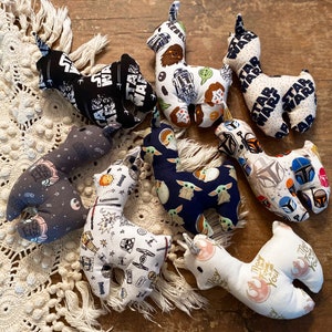 May include: A collection of handmade llama-shaped plush toys with various Star Wars-themed fabric patterns. The toys feature designs with characters, logos, and phrases from the Star Wars franchise. The plushies are arranged on a cream-colored, textured surface.