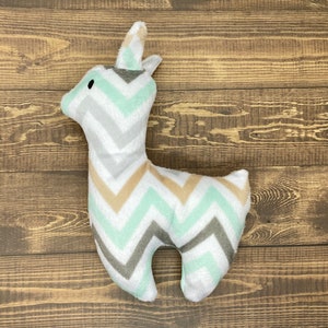 May include: A plush toy shaped like a llama with a chevron pattern in shades of white, gray, and light green. The toy is lying on a wooden surface.