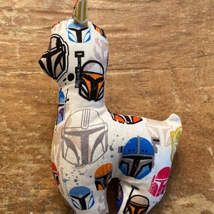 K&ouml;nnte beinhalten: Ein Stofftier-Lama aus wei&szlig;em Stoff, der mit verschiedenen farbenfrohen Mandalorianer-Helm-Designs gemustert ist. Das Lama hat ein kleines, aufrechtes Ohr und steht vor einem Holzgrund.