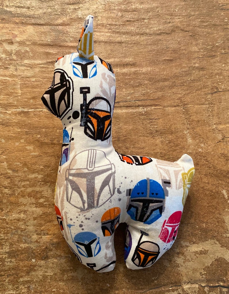 K&ouml;nnte beinhalten: Ein Stofftier-Lama aus wei&szlig;em Stoff, der mit verschiedenen farbenfrohen Mandalorianer-Helm-Designs gemustert ist. Das Lama hat ein kleines, aufrechtes Ohr und steht vor einem Holzgrund.