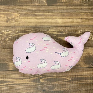 May include: A pink whale-shaped pillow with a white narwhal design and a black eye. The pillow is covered in a white and pink pattern of narwhals and fish.
