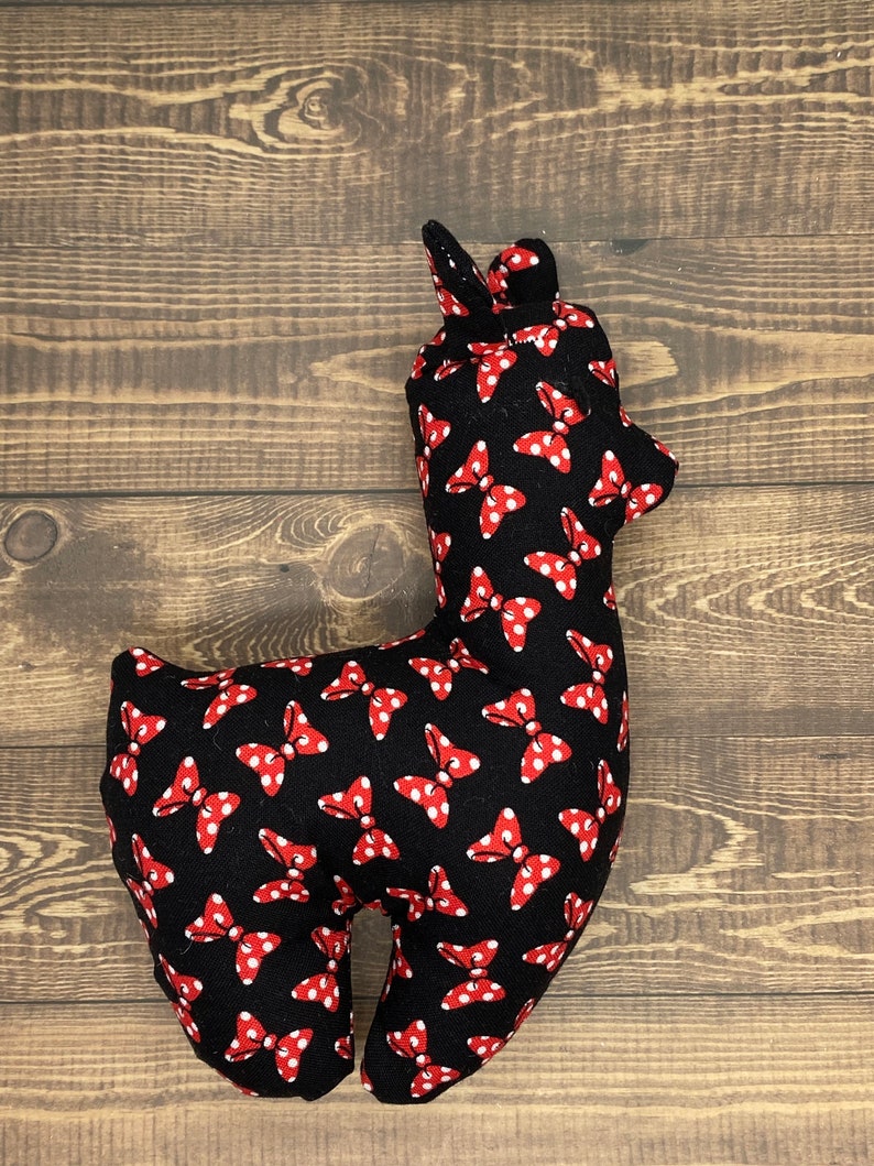 Puede incluir: Un juguete de peluche de alpaca negro con lunares rojos y lazos rojos. La alpaca est&aacute; de pie sobre una superficie de madera.