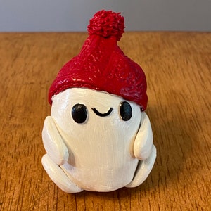 May include: A white clay figurine wearing a red knitted hat with a pom-pom. The figurine has a smiling face with small black eyes and a small mouth.