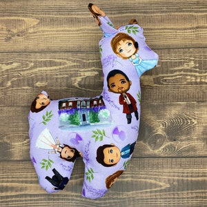 May include: Purple llama-shaped pillow with a whimsical design featuring cartoon characters and a house. The pillow is decorated with lavender flowers and green leaves.