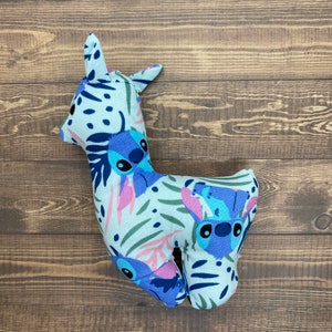 May include: A plush toy shaped like a llama with a white background and blue and purple floral patterns. The llama has a blue and purple Stitch character from the Disney movie Lilo & Stitch on its body.