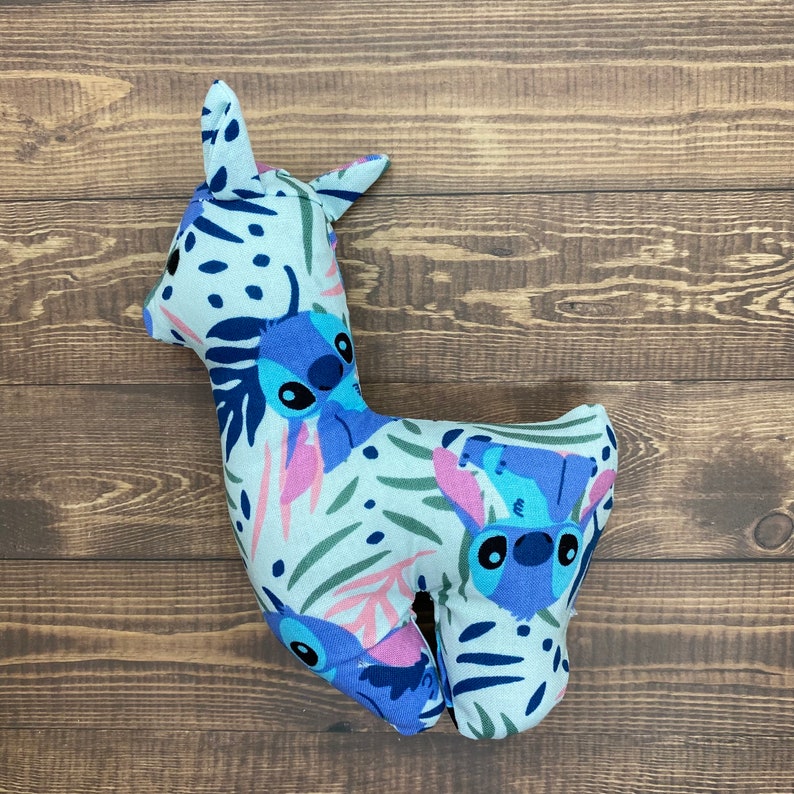 May include: A plush toy shaped like a llama with a white background and blue and purple floral patterns. The llama has a blue and purple Stitch character from the Disney movie Lilo & Stitch on its body.