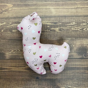 May include: A pink llama-shaped plush toy with a white and pink heart pattern. The toy has a gold glitter accent on the hearts.