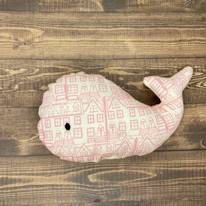 May include: A plush whale toy with a white background and a pink city skyline pattern. The whale has a black eye and a small mouth.