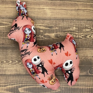 May include: Pink and black plush toy shaped like a llama with a repeating pattern of Jack Skellington and Sally from the Nightmare Before Christmas movie. The pattern also includes hearts and pumpkins. The text "Happy Halloween" is printed on the toy.