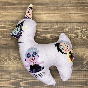 May include: A purple and white pillow shaped like a llama with a cartoon image of Ursula, Maleficent, and Cruella de Vil. The pillow has the word "Ursula" printed on the bottom.