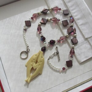 May include: A necklace featuring a pendant of two dolphins, connected by a chain of pearls, square and faceted beads in shades of purple, and clear crystals. The necklace has a silver clasp.