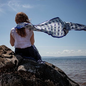 Puede incluir: Una mujer sentada en una roca junto al agua, lleva una blusa blanca y una bufanda con estampado azul y blanco que ondea con el viento.