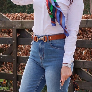 May include: A white long-sleeved button-down shirt with a small dark logo on the chest, paired with blue denim jeans and a tan leather belt featuring a gold-toned metal bit buckle. A colourful patterned silk scarf is tied around the neck.