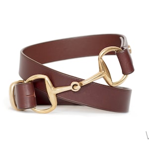 Peut inclure: Une ceinture en cuir marron foncé dotée d'un mors en métal doré poli. La ceinture est enroulée, mettant en valeur la texture lisse de la lanière en cuir et les détails métalliques. Le nom de la marque VEZAVI est imprimé dans le coin inférieur droit.