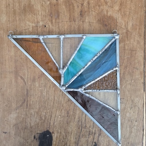 Peut inclure: Un attrape-soleil en vitrail de forme triangulaire. Il présente des sections de verre marron, beige, turquoise, bleu et transparent, encadrées d'une bordure en métal argenté. Deux petites boucles sont en haut pour l'accrocher.