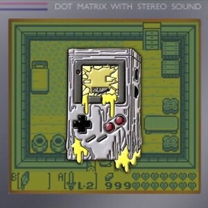 May include: A silver and yellow enamel pin of a retro handheld video game console with a melted appearance. The pin has a pixelated screen with a yellow and white image inside. The pin also has a D-pad, two buttons, and a text display that reads "L-21 999".